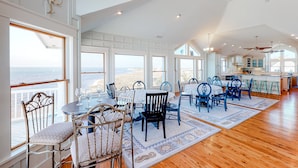 A spacious dining area with multiple tables and chairs, large windows offering ocean views, and a modern kitchen in the background. - Waterfront home with amazing sound view, private pool, hot tub, & game room (Avon)