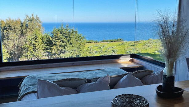 A room with a large window overlooking a coastal landscape, a wooden table, and a vase with dried plants. - Chackra Centinela  (Navidad)