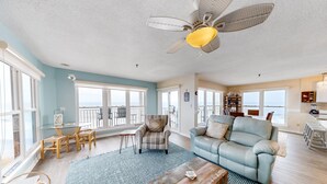 Un salon spacieux doté d’un grand ventilateur de plafond, d’un fauteuil bleu, d’une table basse en bois et offrant une vue sur l’océan grâce à de grandes fenêtres.