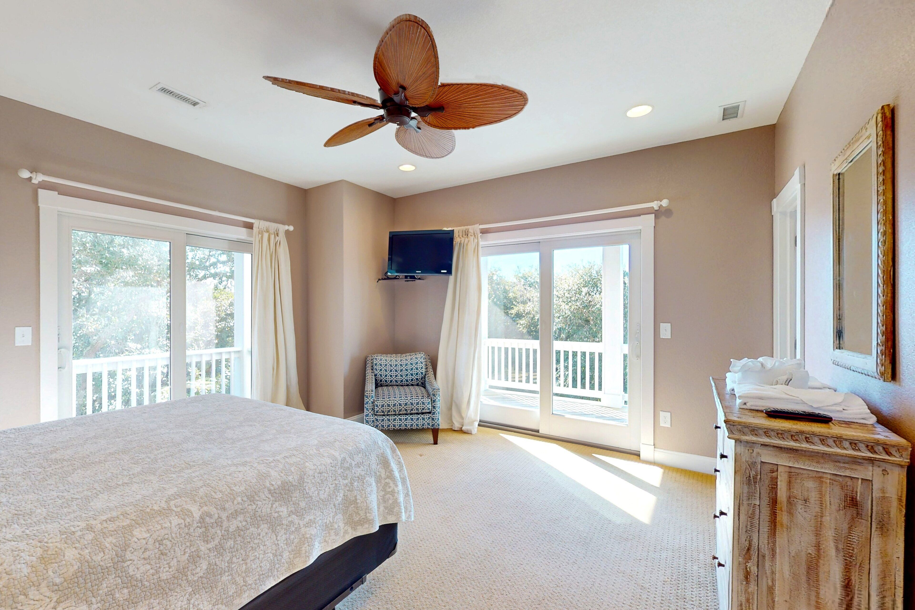 A bedroom with a bed, a ceiling fan, a TV, a dresser, and large windows.