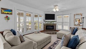A living room with a fireplace, two sofas, a flat-screen TV, and large windows with blinds. - Fore Buoys: Canalfront, dock, close to beach! (Ocean Isle Beach)
