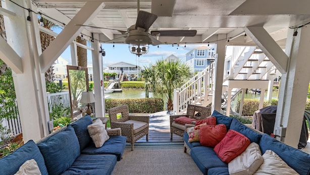 A covered outdoor seating area with blue sofas and wicker chairs, a ceiling fan, and a view of a residential neighborhood. - Fore Buoys: Canalfront, dock, close to beach! (Ocean Isle Beach)