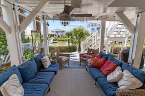 A covered outdoor seating area with blue sofas and wicker chairs, a ceiling fan, and a view of a residential neighborhood. - Fore Buoys: Canalfront, dock, close to beach! (Ocean Isle Beach)