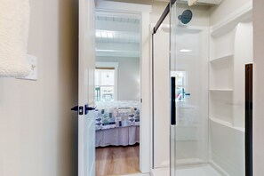 Ein Badezimmer mit Duschkabine aus Glas, einem weißen Waschbecken und einer sichtbaren Tür, die in ein Schlafzimmer mit Bett und Fenster führt.