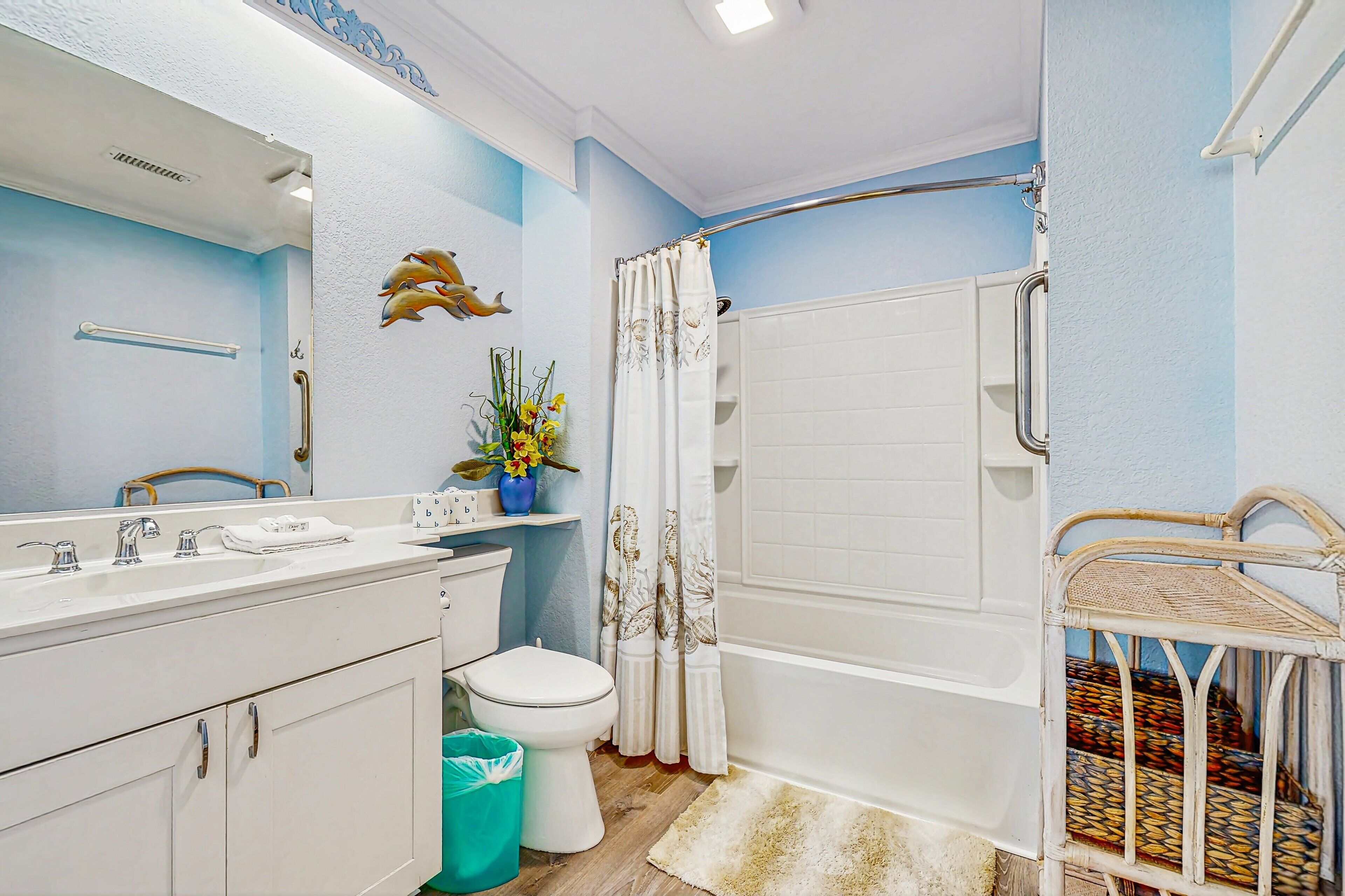 Une salle de bain avec un lavabo, des toilettes et une baignoire, tous de couleur blanche. On y trouve un rideau de douche à motifs floraux, une poubelle blanche et une chaise en osier.