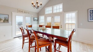 A spacious dining area with a glass-top table surrounded by wooden chairs, a chandelier, and a view of the beach through large windows. - Dog-friendly oceanfront home with private outdoor pool, hot tub & central AC (Waves)