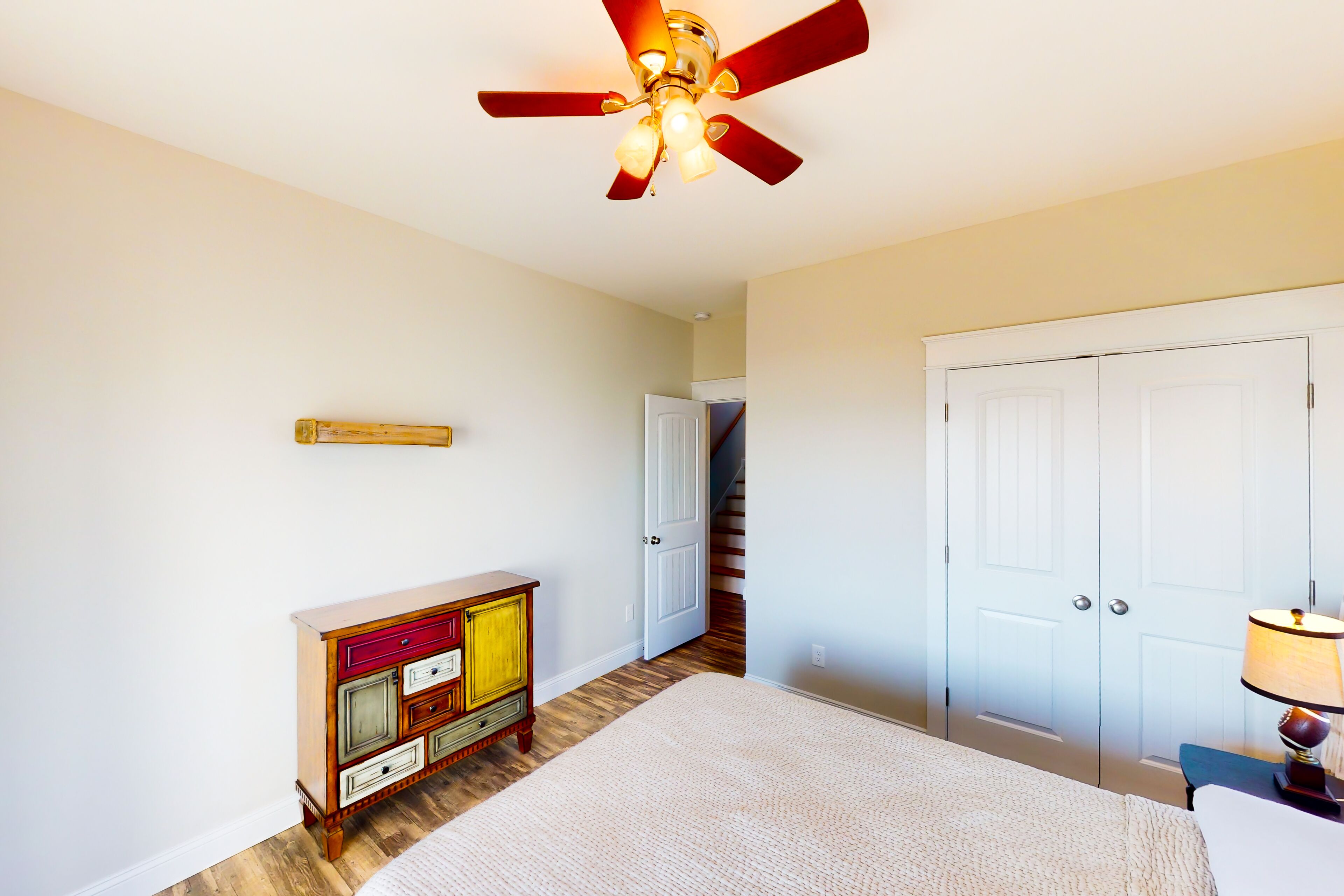 A bedroom with a bed, a ceiling fan, a dresser, a door, and a lamp.