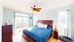 A bedroom with a large bed, a ceiling fan, a nightstand, a dresser, and a view of the ocean through a sliding door. - Beautiful dog-friendly ocean view home with hot tub, lovely balcony, W/D & AC (North Topsail Beach)