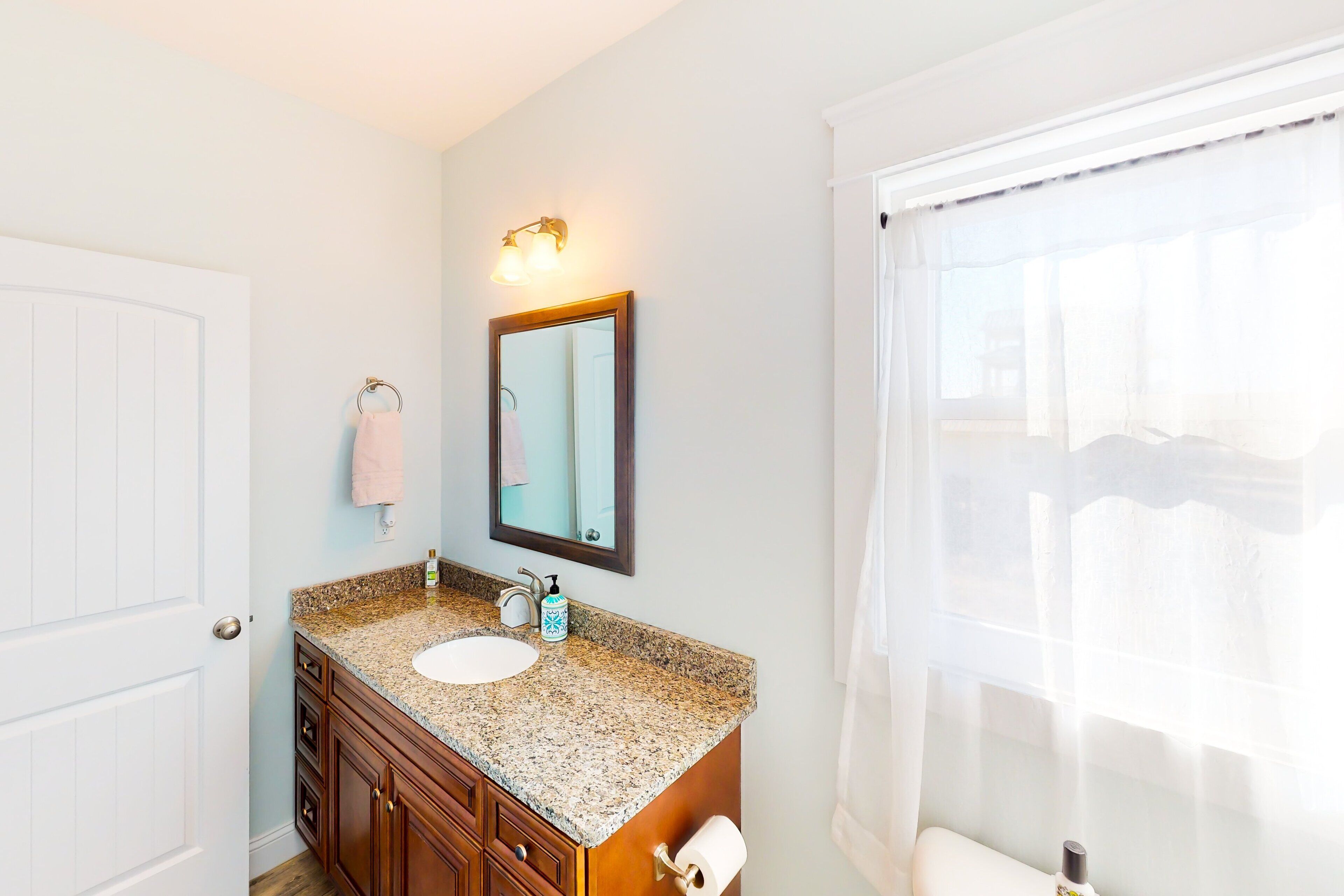 Une salle de bain avec une porte blanche, un lavabo sur un plan de travail en granit, un miroir et une fenêtre ornée de rideaux légers.