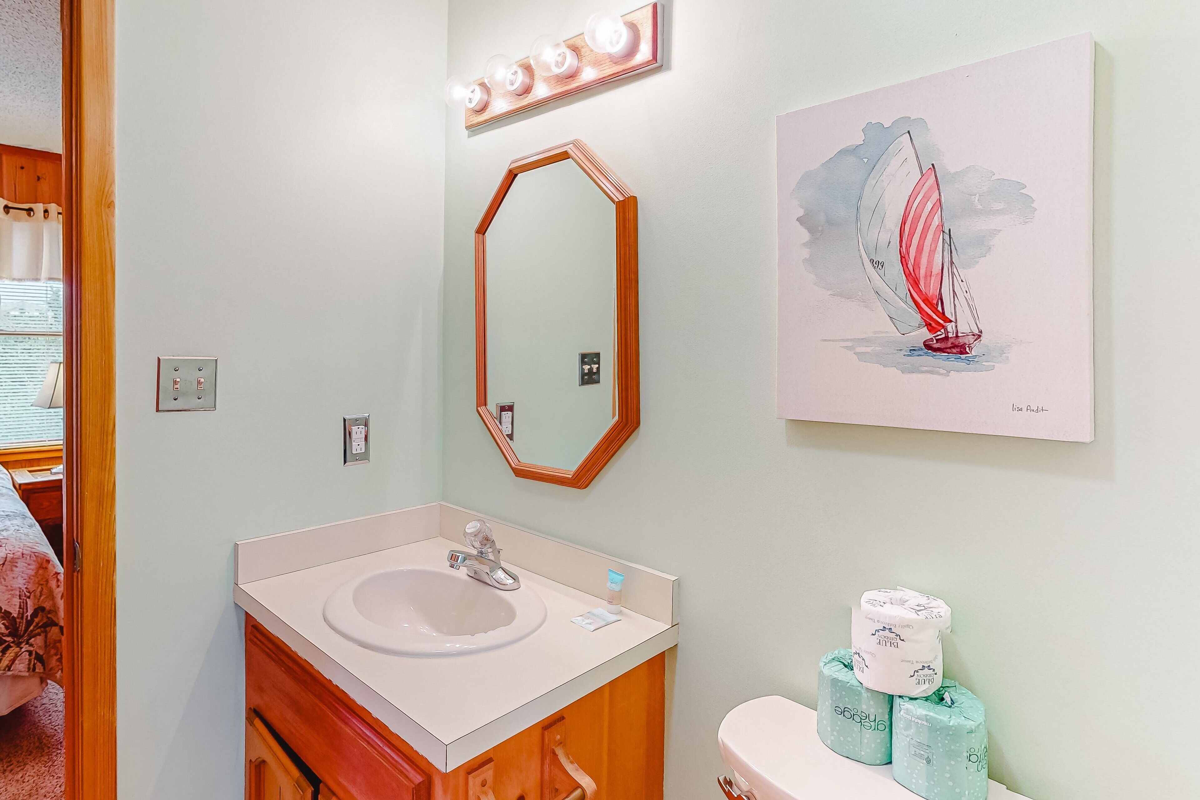 A bathroom with a sink, mirror, and toilet. There is a framed artwork of a sailboat on the wall.