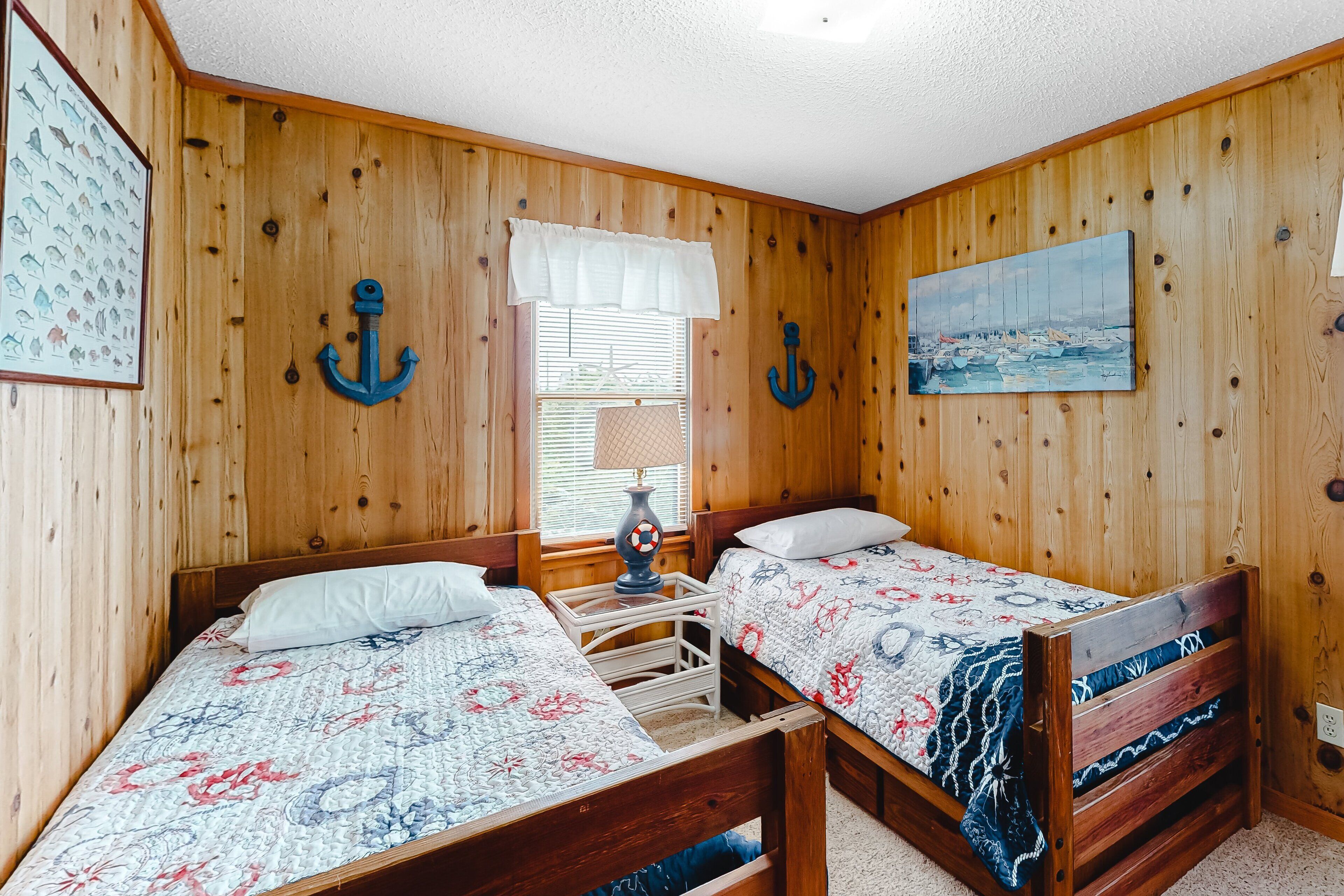 A room with wooden walls, two beds with floral bedding, a window with blinds, and a painting of a seascape.