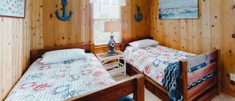 A room with wooden walls, two beds with floral bedding, a window with blinds, and a painting of a seascape.