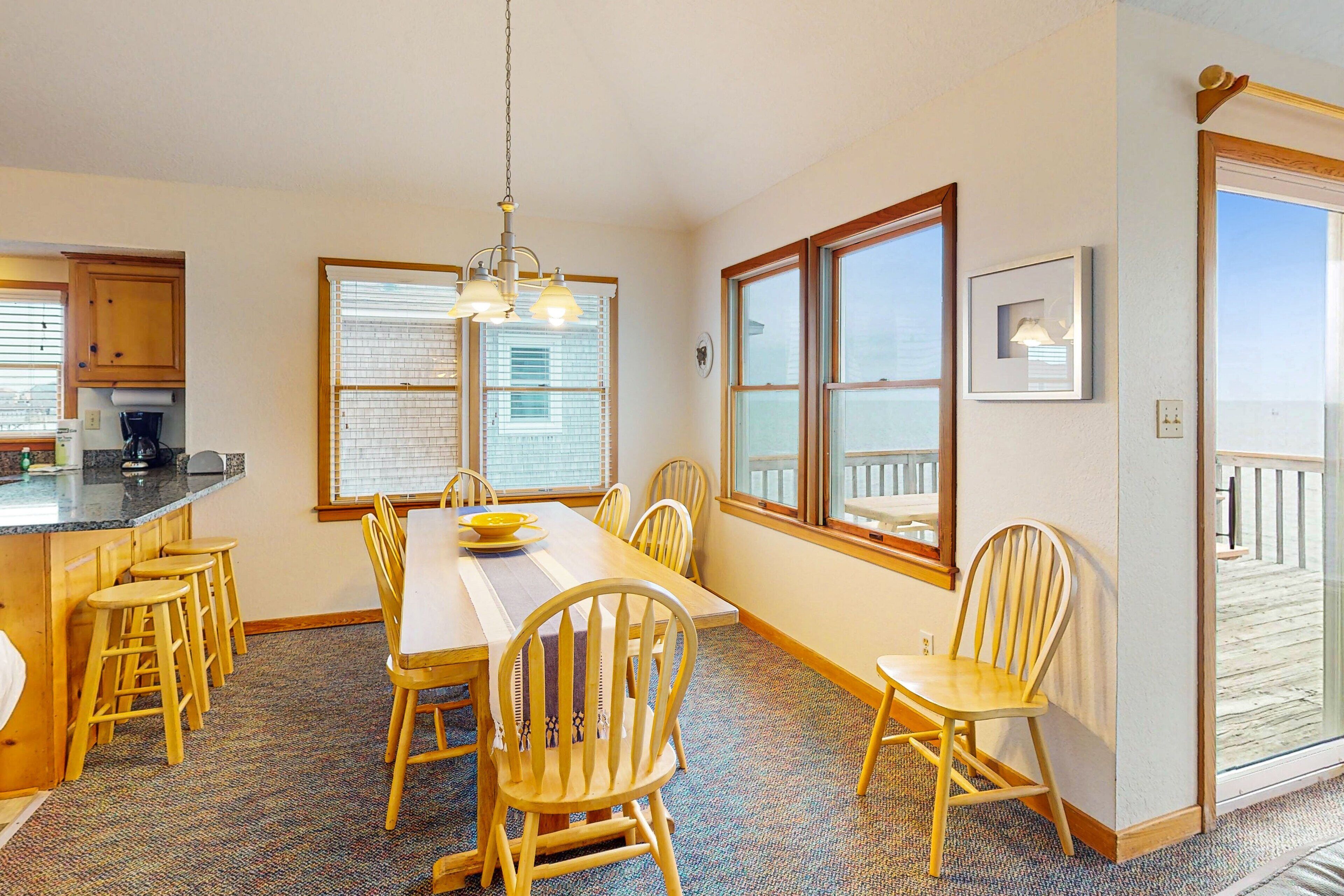 A dining area with a long table, wooden chairs, and a chandelier. There are windows with blinds and a view of the outside.
