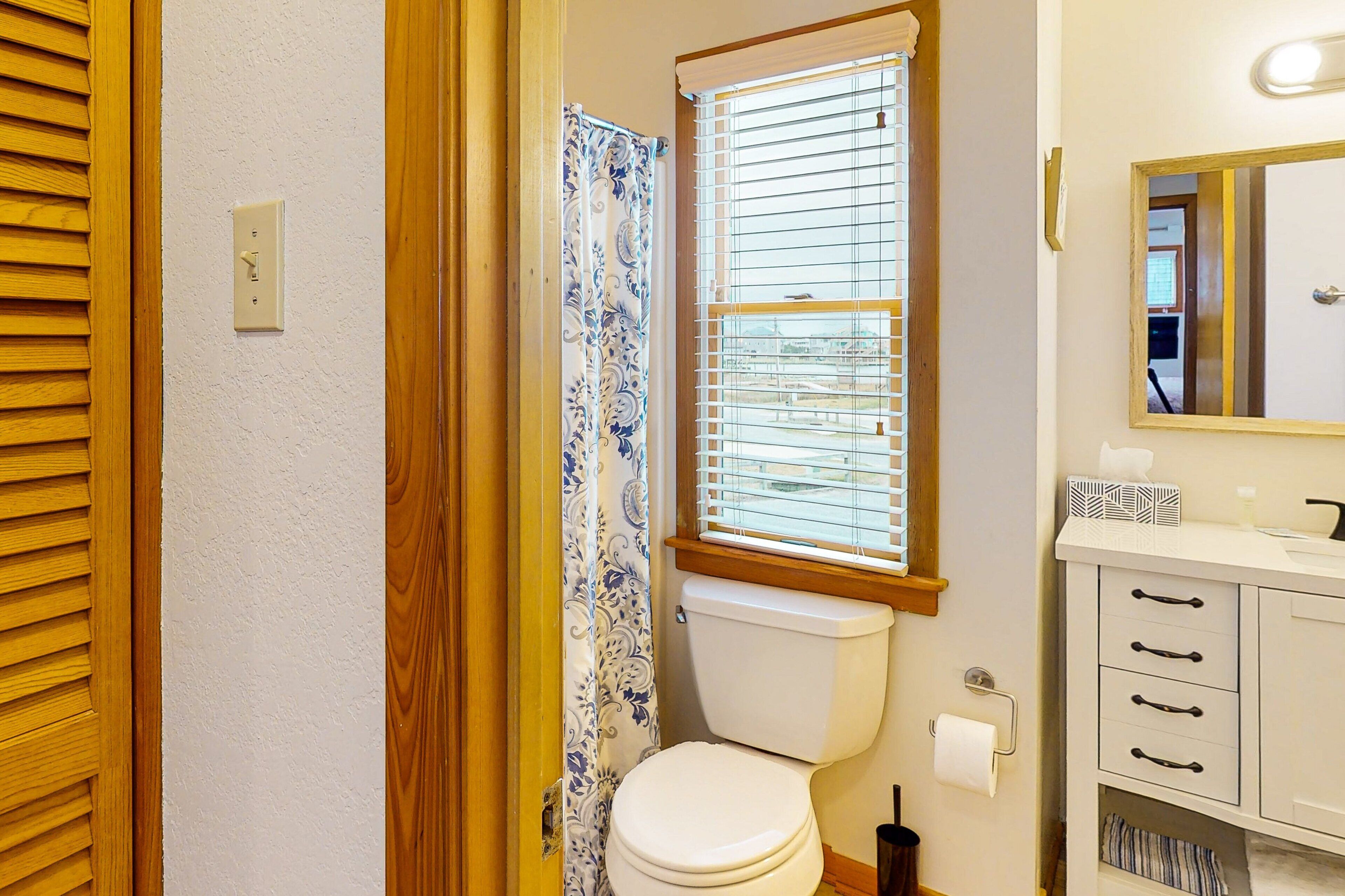 Un baño con inodoro, ventana con persianas, cortina de ducha y tocador con cajones.