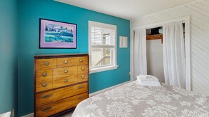Quarto com paredes verde-água, cómoda de madeira com gavetas, cama com colcha floral, janela com portão branco e quadro pendurado na parede.