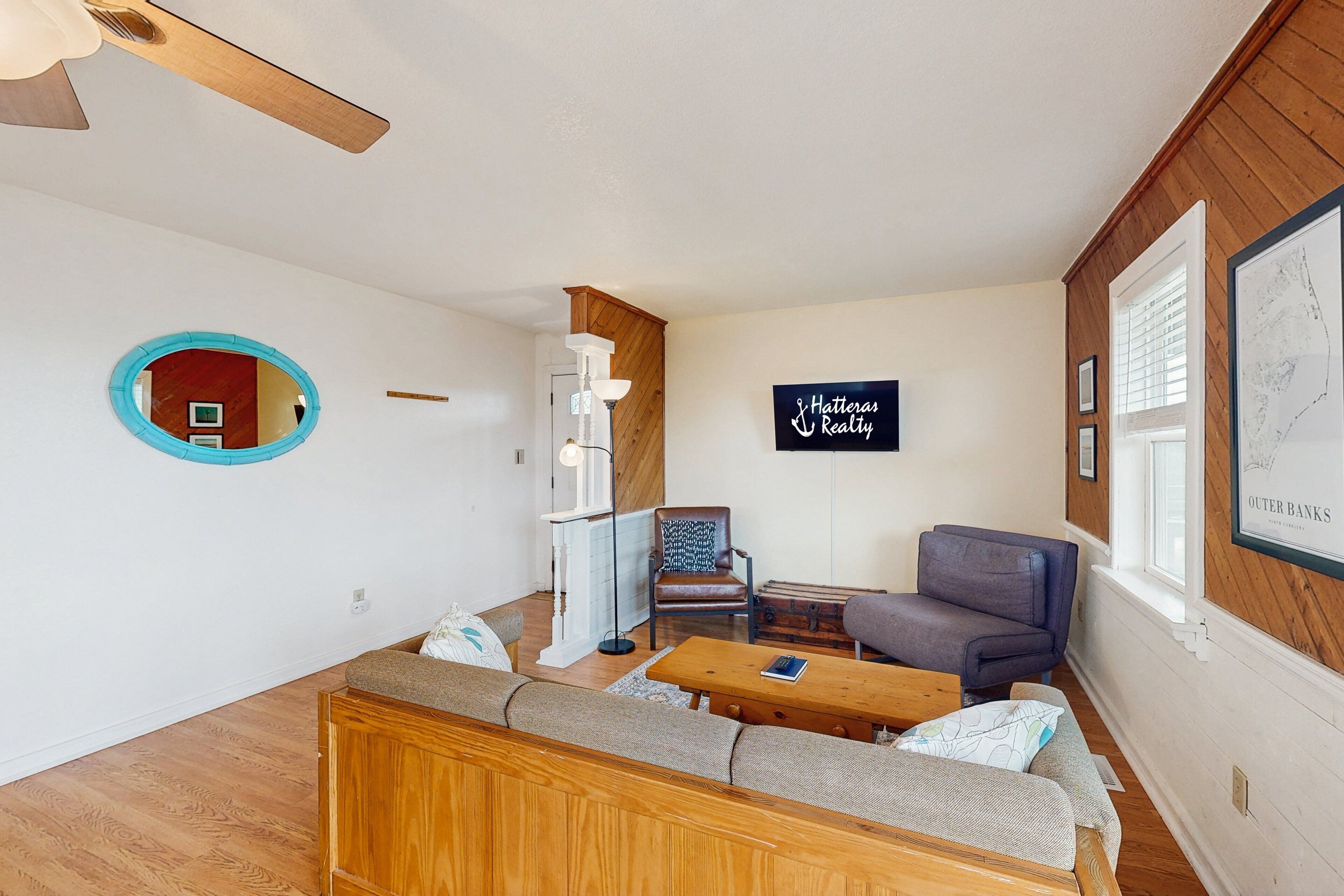Una sala de estar moderna con ventilador de techo de madera, un espejo redondo, un sofá, una mesa de centro y un cartel que dice 'Hatteras Realty'.