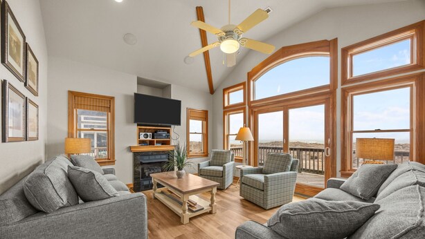 Ein großzügiges Wohnzimmer mit grauen Sofas, einem Holztisch und einem Deckenventilator. Große Fenster bieten einen Blick ins Freie.