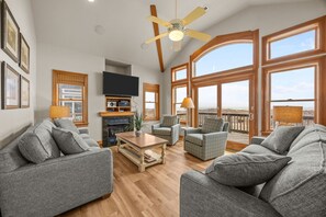 A spacious living room with grey sofas, a wooden coffee table, and a ceiling fan. There are large windows offering a view of the outdoors.