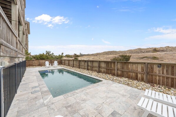 A swimming pool with a wooden fence, stone tiles, and white chairs.
