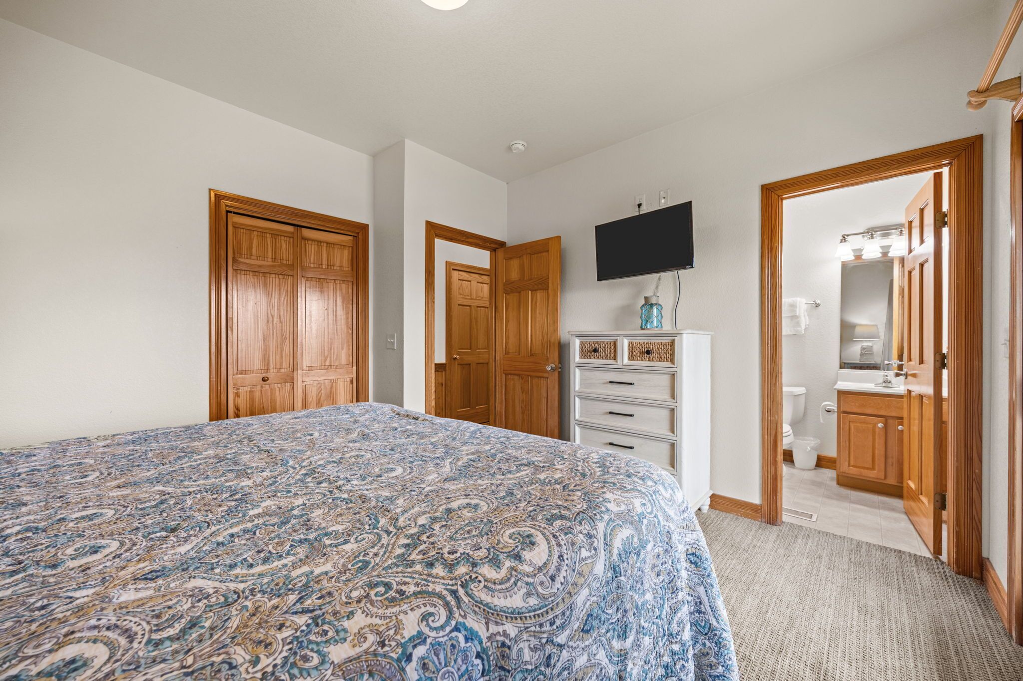 Un dormitorio con una cama grande, un armario de madera, un televisor montado en la pared y un baño visible a través de una puerta abierta.