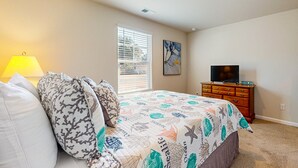 A bedroom with a bed, a dresser, a television, a window with blinds, and a lamp. - Salty Retreat: Fire pit | close to golf courses (Ocean Isle Beach)