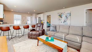 A modern living room with a sofa, a coffee table, and a leather chair. There is a kitchen area with bar stools and a refrigerator. - Salty Retreat: Fire pit | close to golf courses (Ocean Isle Beach)