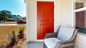 A red door, a wicker chair, and a window with blinds. - Salty Retreat: Fire pit | close to golf courses (Ocean Isle Beach)
