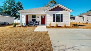 A single-story house with a red door, a front porch with two chairs, and a small garden area. - Salty Retreat: Fire pit | close to golf courses (Ocean Isle Beach)