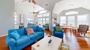 Un salón espacioso con un sofá azul, una mesa de centro blanca y una silla de madera. Hay ventiladores de techo, ventanas amplias y vistas al exterior.