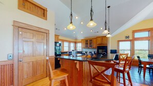 A spacious kitchen with wooden cabinets, a large island with bar stools, pendant lighting, and large windows offering a view. - Dog-friendly ocean- & sound-view home with private pool, hot tub, & central AC (Avon)