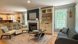 A modern kitchen with white cabinets, a dining area with bar stools, a fireplace, and a living room with a sofa and coffee table.