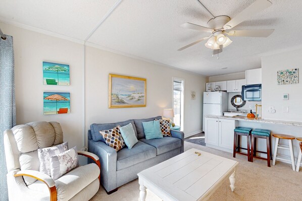 A living room with a sofa, armchair, and coffee table. There is a kitchen area with a refrigerator, microwave, and sink. - Cheerful beachfront condo with balcony, tennis court, & W/D - snowbird-friendly (North Topsail Beach)