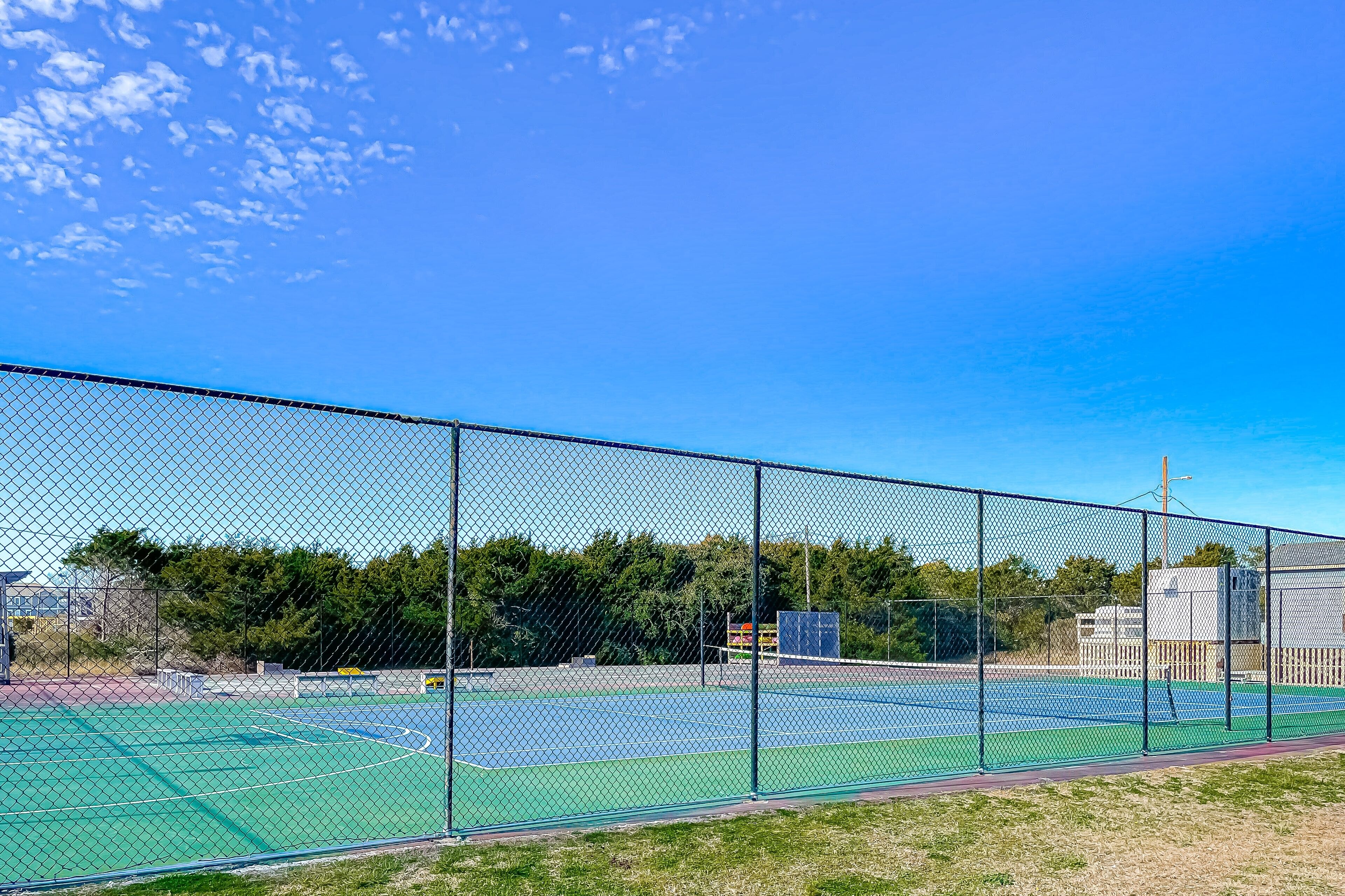 Un campo da tennis all'aperto circondato da una rete metallica, con un cielo azzurro e alberi sullo sfondo.