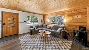 A cozy cabin interior with a wooden ceiling, a sofa set, a fireplace, and large windows.