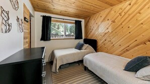 A small, cozy room with wooden ceiling and walls, two single beds, a desk, and a window with curtains.