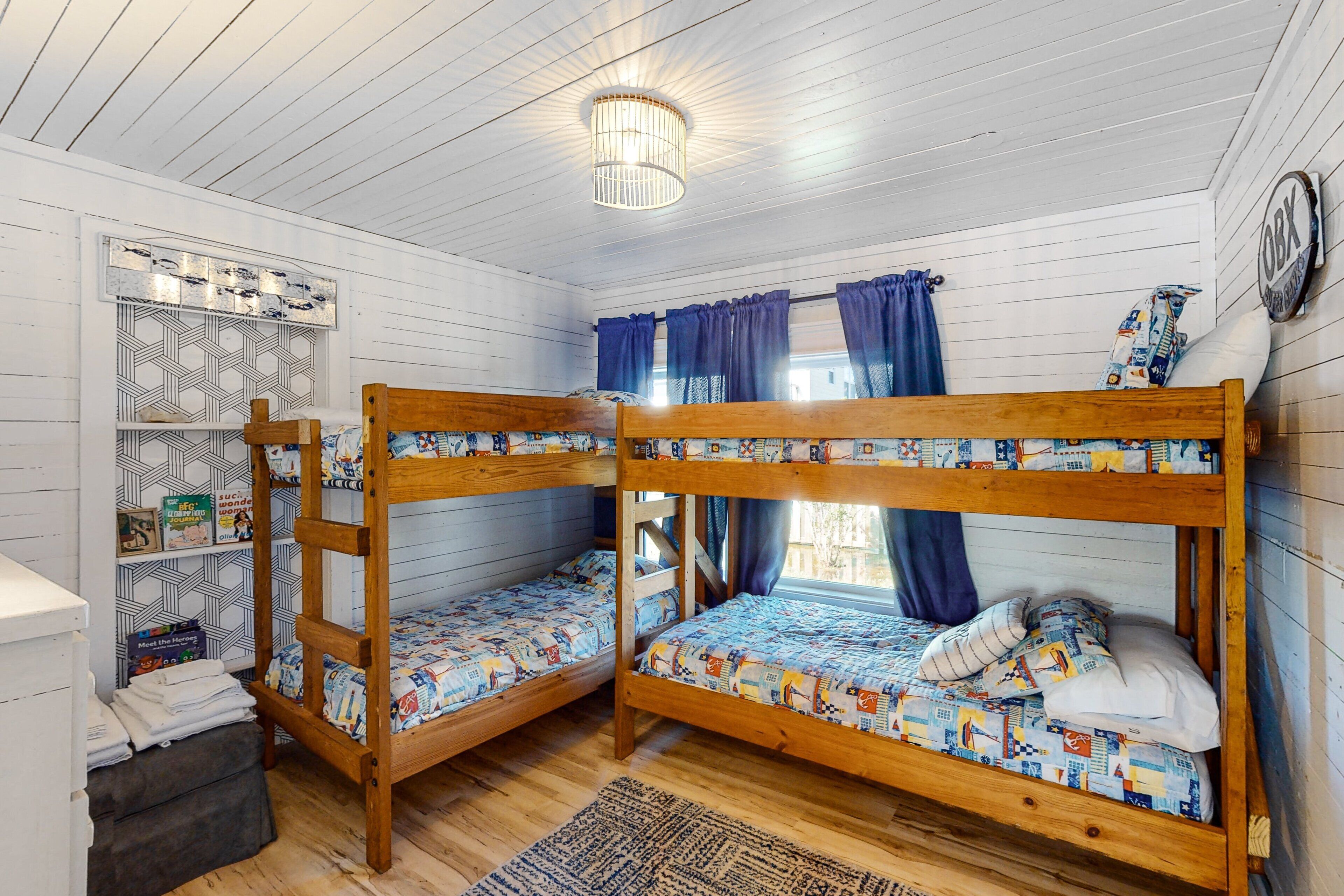 A bunk bed room with a wooden ceiling, a small shelf with books, and a window with curtains.