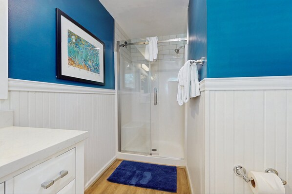 Een badkamer met een glazen douchecabine, een wit wastafeltje, een kunstwerk in een lijst aan de muur en een blauwe badmat.