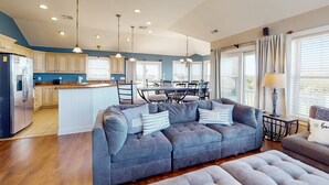 A modern kitchen with a built-in island, a dining area with a table and chairs, and a living room with a grey sofa and ottoman.