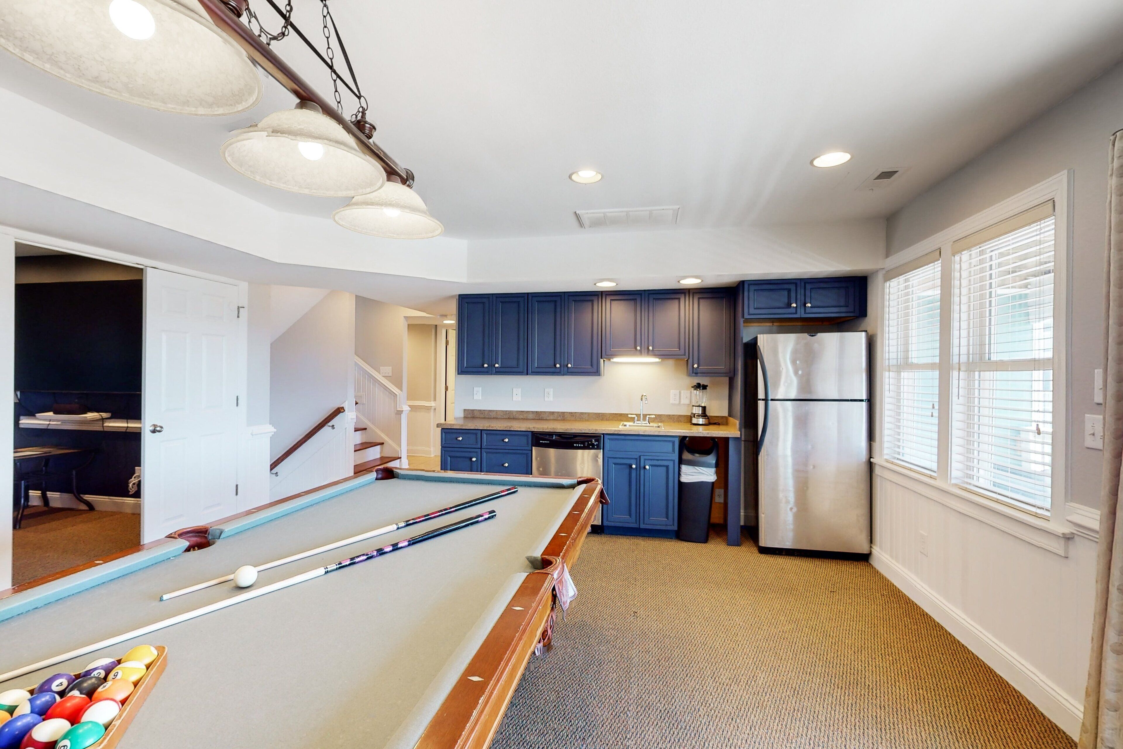 A pool table in a room with a kitchen area, a refrigerator, and a staircase leading to another room.