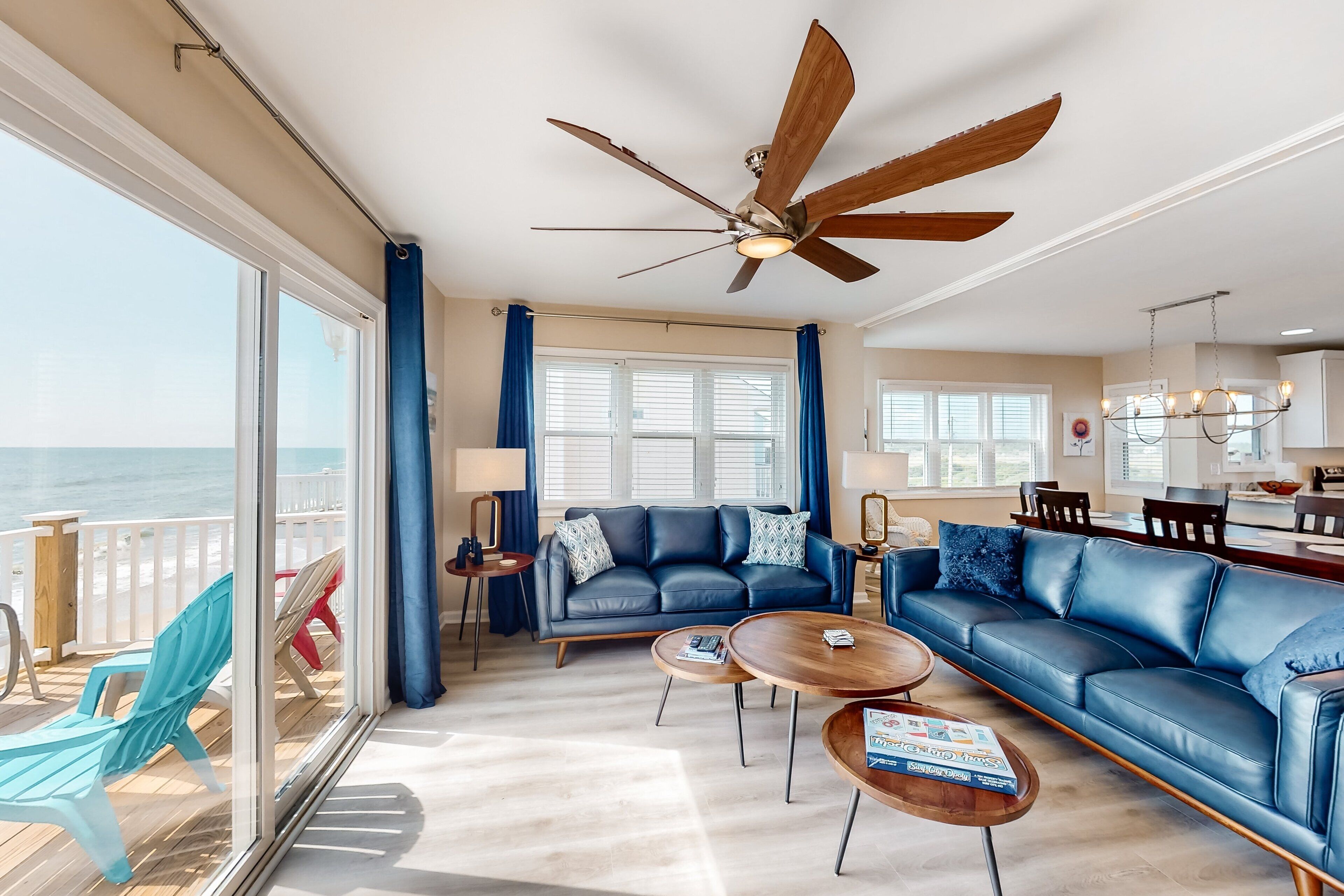 A spacious living room with a large ceiling fan, a blue sofa, a coffee table, and a view of the ocean through large windows.