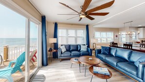 A spacious living room with a large window overlooking the ocean, a ceiling fan, and a dining area with a chandelier. - Renovated townhome right on the ocean with private beach & coffee stations (North Topsail Beach)