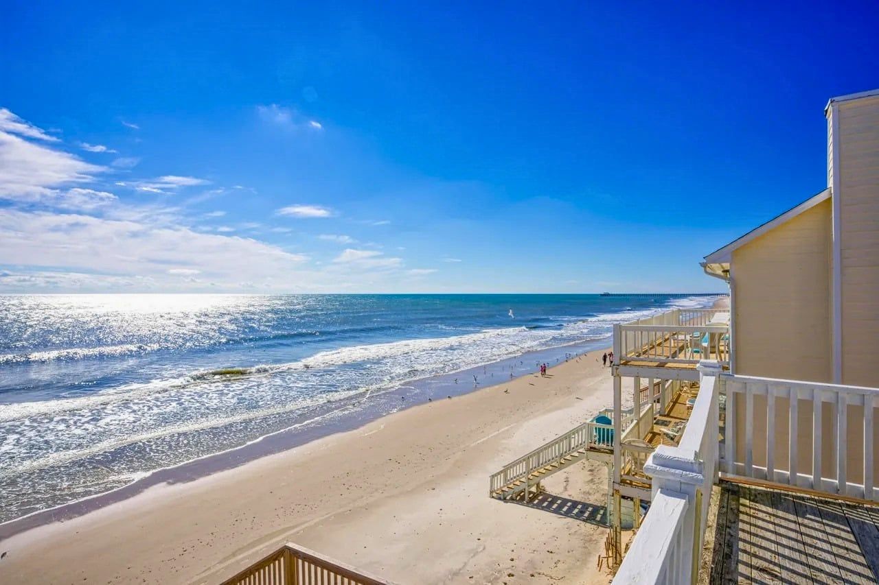 A beachfront hotel with a balcony overlooking the ocean, sandy beach, and a clear sky.