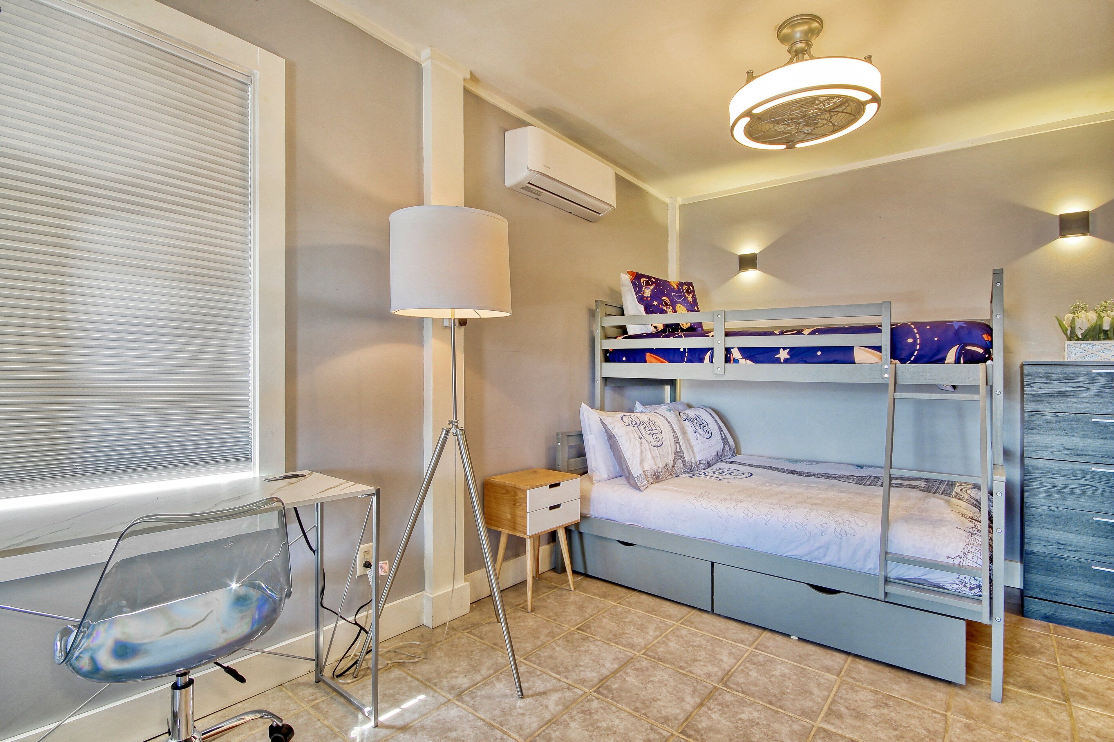 Une chambre avec un lit superposé, un bureau avec une chaise, une lampe et une fenêtre avec des stores.