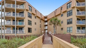 A wooden walkway leading to a multi-story building with balconies and a palm tree. - Charming ocean-view condo with washer/dryer, on-site pool (North Topsail Beach)