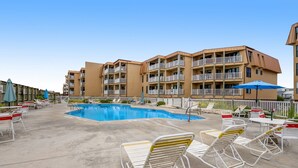 A hotel pool area with multiple lounge chairs and umbrellas, surrounded by a building with balconies. - Charming ocean-view condo with washer/dryer, on-site pool (North Topsail Beach)