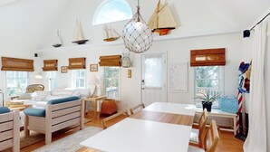 Ein geräumiges Wohnzimmer mit einem großen weißen Tisch, Holzstühlen, einem blauen Sofa mit Kissen und einer hängenden Lampenleuchte.