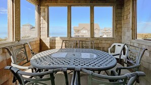 A covered patio with a round table and chairs, large windows, and a view of beachfront houses. - 3-story Pet friendly home with private outdoor pool, hot tub, pool table, & elevator (Waves)