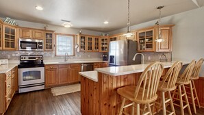 A kitchen with wooden cabinets, a stainless steel refrigerator, a microwave, and a stove. There are wooden bar stools at the island. - 3-story Pet friendly home with private outdoor pool, hot tub, pool table, & elevator (Waves)