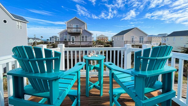 Une terrasse dotée de chaises Adirondack turquoise et d’une petite table, offrant une vue sur un quartier résidentiel avec ses maisons et un ciel bleu dégagé.