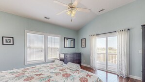 Une chambre avec un grand lit, un ventilateur de plafond, une commode et une porte-fenêtre coulissante donnant sur un balcon.